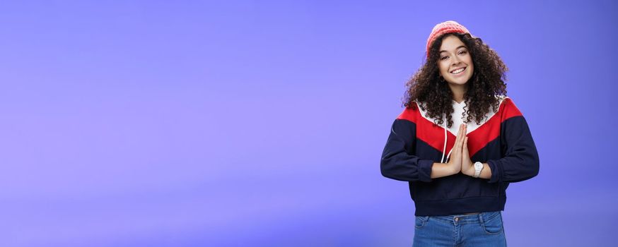 Portrait of charming curly-haired feminine girlfriend begging for favour as posing over blue background in winter outfit with hat, holding hands in pray smiling with angel look and friendly gaze. Body language and facial expressions concept