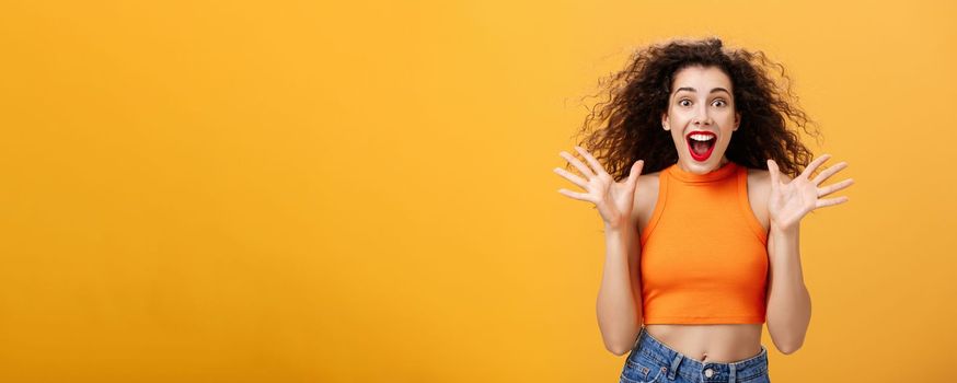 Excited impressed attractive stylish woman with nose ring, red lipstick and curly hairstyle smiling amazed with opened mouth gesturing with raised palms being astonished over orange background. Emotions concept