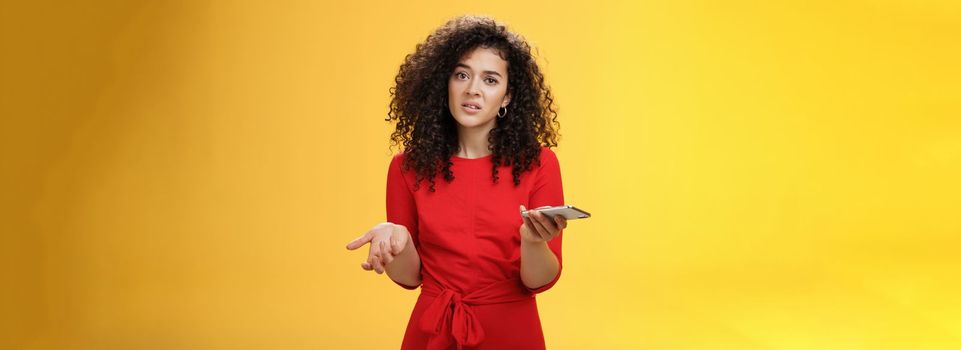 So waht, I confused. Questioned uncertain woman with curly hair in red dress shrugging looking clueless as holding hand cannot understand where order as checking mail box via device over yellow wall. Technology and internet concept