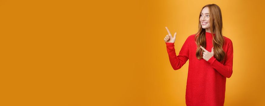 Interested gentle and tender young 20s ginger woman in red warm sweater pointing, looking at upper left corner with broad smile, enjoying nice happy scene over orange background. Advertisement concept