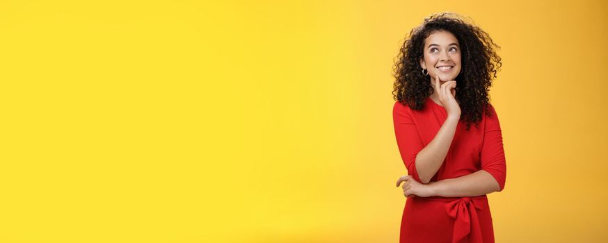 Girl having interesting idea, smiling as feeling confident plan might work. Charming happy woman with curly hair in red dress gazing at upper left corner thoughtful, thinking over yellow wall.