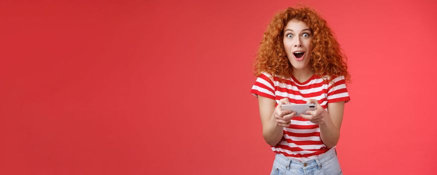 Lifestyle. Impressed excited good-looking redhead curly woman hold smartphone horizontal open mouth fascinated stare camera thrilled astonished win passed level game happy playing cool app red background.