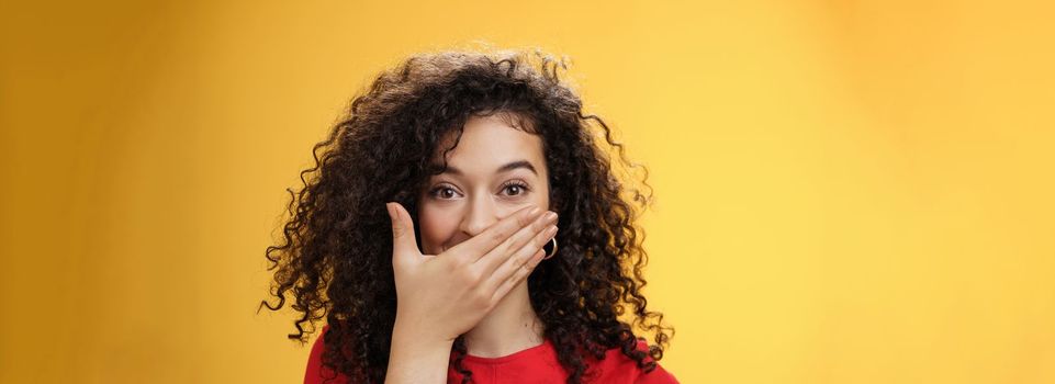 Close-up shot of excited and happy attractive female with curly hair giggling, chuckling and covering mouth as smiling standing amused and joyful as mocking friend over yellow background. Emotions and facial expressions concept