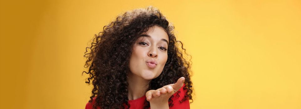 Sensual flirty and tender attractive girlfriend with curly hairstyle folding lips extending hand to send air kiss at camera, smiling flirting and being coquettish over yellow background. Romance, people and relationship concept