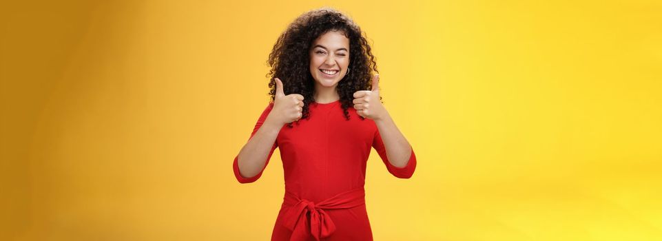 Creative and charismatic happy upbeat woman 25s with curly hair in red dress winking in approval and showing thumbs up with broad smile, satisfied giving positive reply over yellow wall.