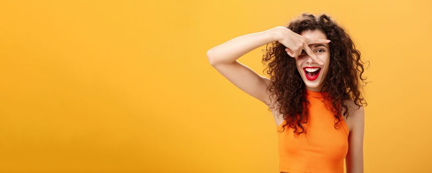 Waist-up shot of creative joyful caucasian female. with curly hairstyle and small tattoo showing peace gesture and peeking through fingers at camera happily smiling broadly having fun over orange wall.