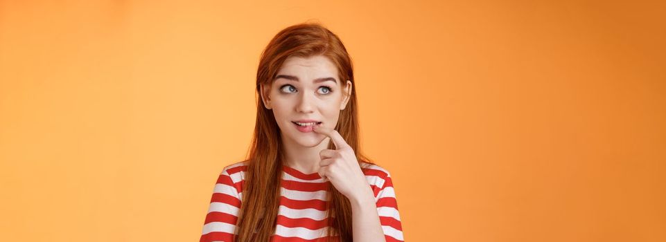 Curious sly cute redhead girlfriend, thinking silly biting finger, look interested upper left corner, have inriguing idea, ponder choice, deciding order online, stand thoughtful orange background.