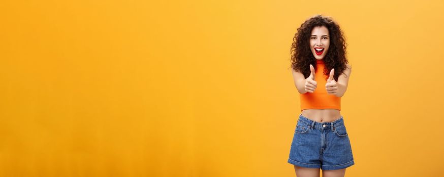 Charismatic ambitious and outgoing charming caucasian. woman with curly hairstyle and red lipstick showing thumbs up gesture in like or approval smiling joyfully being supportive over orange wall.