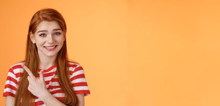 Close-up silly cute redhead female trying help showing way, smiling friendly politely introduce promo, pointing upper left corner smiling modest, stand orange background give advice.