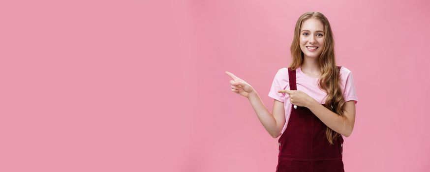 Lifestyle. Polite and charismatic pretty young girl with long fair hair in corduroy trendy overalls pointing left as if inviting or showing something interesting smiling friendly at camera over pink background.