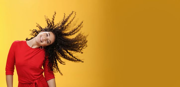 Girl getting carefree and wild waving hair and making sun with curls flying in air smiling broadly tilting head and having fun, dancing delighted and amused feeling playful against yellow background.