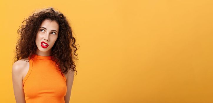 Stylish hot dark-haired woman with curls and red lipstick. in trendy cropped top looking at upper left corner intrigued and curious clenching teeth sensually posing over orange background.