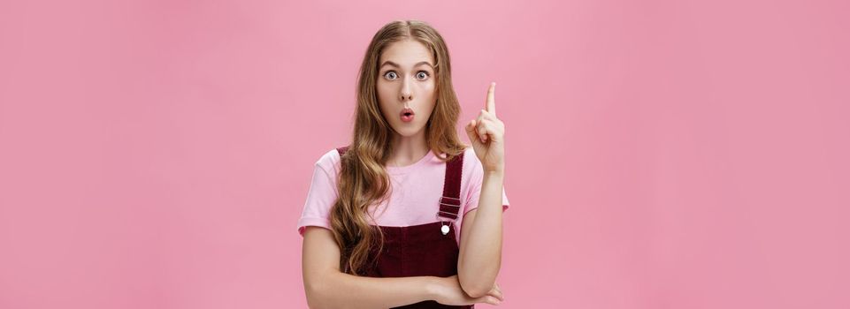 Young smart and creative female student sharing with team awesome idea coming in mind raising index finger in eureka gesture looking excited at camera folding lips saying suggestion over pink wall. Body language, body-positive and gestures concept