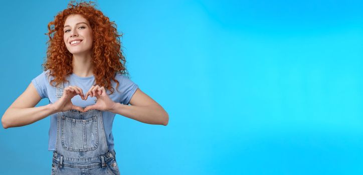 Sassy confident good-looking modern redhead curly woman raise head proud love tell girlfriend romantic heartwarming feelings present own heart smiling broadly standing blue background.