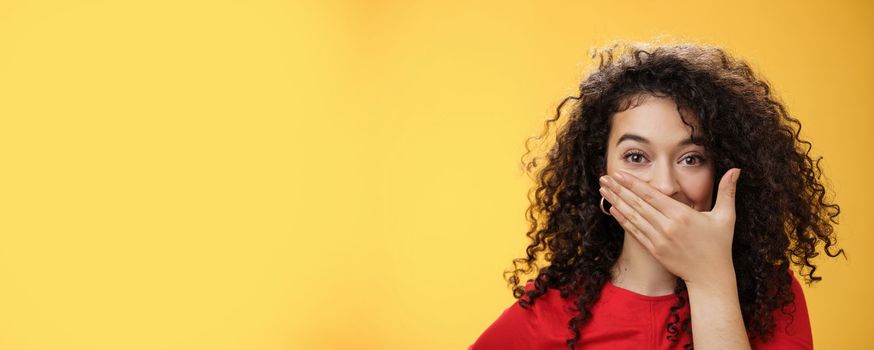 Close-up shot of excited and happy attractive female with curly hair giggling, chuckling and covering mouth as smiling standing amused and joyful as mocking friend over yellow background. Emotions and facial expressions concept