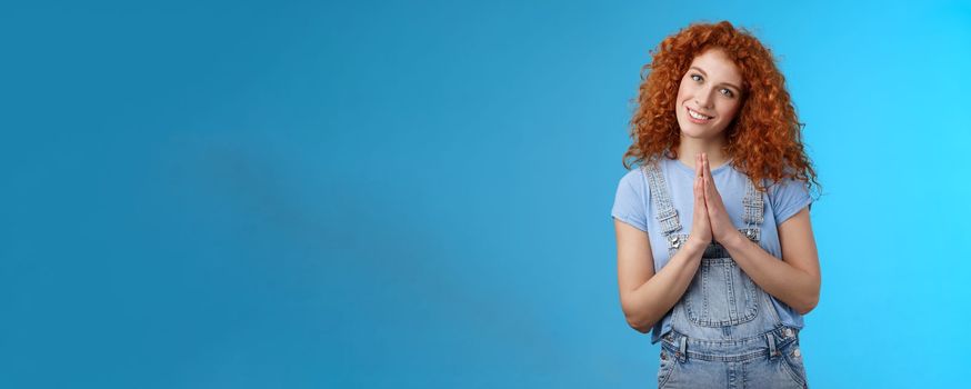 Lifestyle. Girl acting like nun redhead cute curly female press palms together pray supplication gesture tilting head silly smiling innocent angel gaze pleading begging favour asking something blue background.