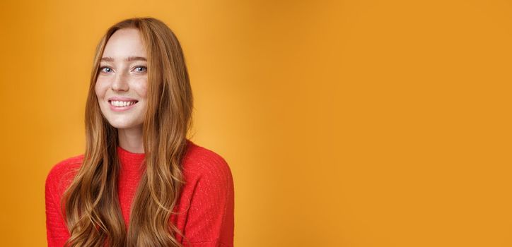 Close-up shot of happy charismatic and carefree cute redhead girl in red knittes outfit smiling joyfully as turning at camera satisfied and joyful posing delighted over orange background. Facial expressions and emotions concept