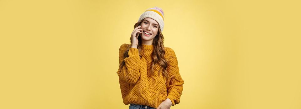 Charming stylish european woman wearing hat sweater tilting head flirty smiling talking smartphone calling boyfriend consulting friend via cellphone, standing happily casual pose yellow background.