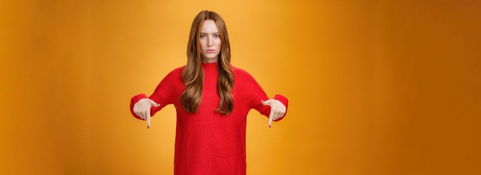 Displeased serious-looking female in knitted red dress frowning looking concerned as pointing down at displeasing unappropriate thing standing disappointed and unhappy over orange background.