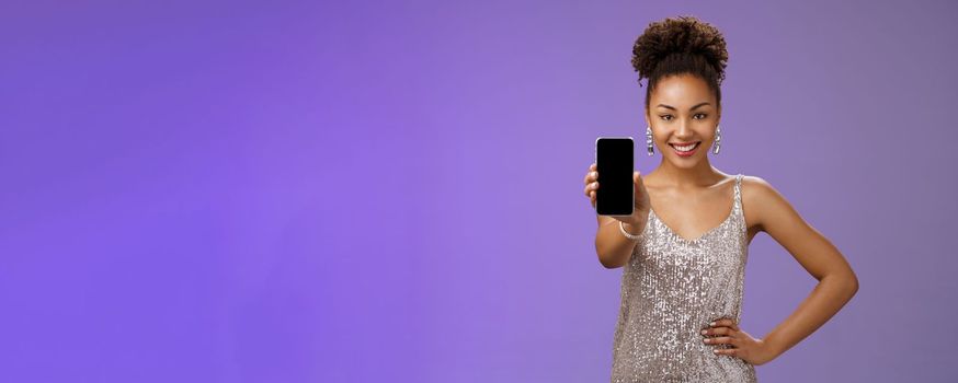 Sassy elegant african-american woman. in glittering shiny silver dress hold hand waist confident pose gladly smiling extend arm showing smartphone display check out cool app blue background.