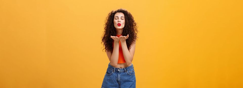 Portrait of sensual and joyful charming tender woman with curly hairstyle red lipstick and cropped top closing eyes folding lips in mwah bending towards opened palms to blow air kiss at camera. Relationship and emotions concept