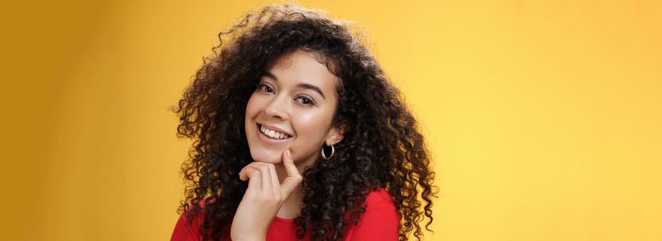 Lifestyle. Headshot of cute silly and tender feminine romantic woman with curly hairstyle touching lip with index finger making eyes at camera and smiling as using seduction skills over yellow background.