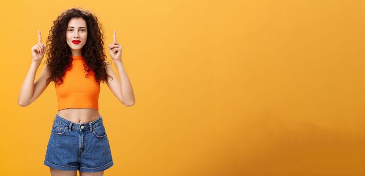 Portrait of confident charismatic caucasian female. with curly hairstyle in red lipstick and orange cropped top pointing up with raised hands and smiling cheerfully at camera assured copy space fine.