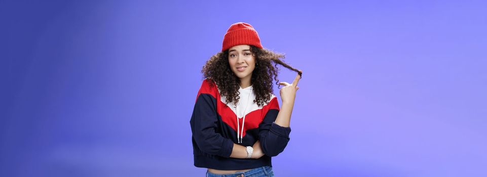 Stylish and snobbish arrogant curly-haired woman in warm beanie rolling curl on finger smirking and looking with contempt at camera, scorning person as being too cool over blue background.