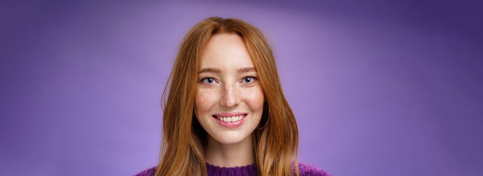 Headshot of charming attractive and happy young redhead woman with freckles and bright white smile, grinning satisfied at camera as posing over purple background friendly and delighted.