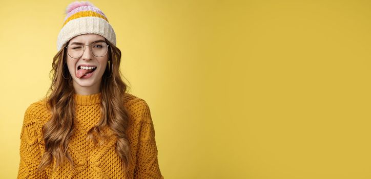 Carefree attractive funny happy caucasian girl 20s curly-haired wear glasses hat sweater having fun playfully show tongue, winking flirty expressing positive optimistic mood, yellow background.