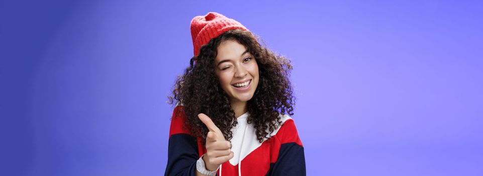 Lifestyle. Friendly-looking sociable and stylish woman with curly hair in red beanie winking joyfully and pointing with finger pistol at camera as greeting mate being cool and confident over blue wall.