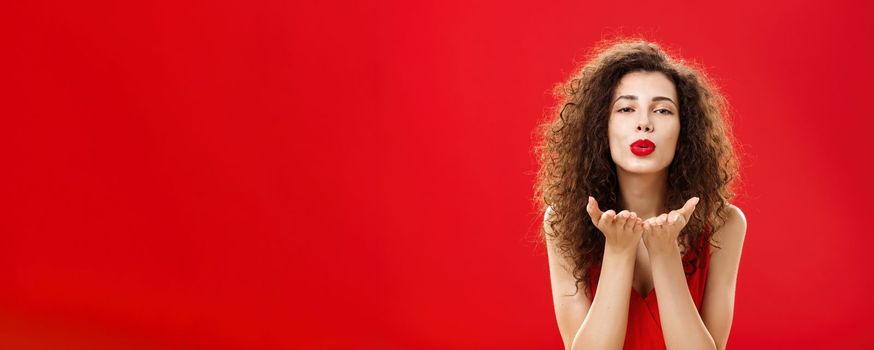 Woman rewards boyfriend with passionate wind kiss after romantic date. Portrait of sensual hot adult female in red elegant dress with curly hairstyle bending towards camera sending mwah flirty. Romance concept