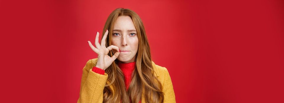 Girl putting seal on mouth making promise not tell anyone secret, sucking lips and holding finger near as zipping not spill words, looking serious and determined to keep surprise safe over red wall.