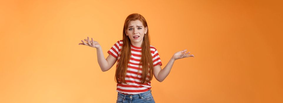 Careless arrogant redhead rich girl say so what, not giving care what people thing, look dismay ignorant shrugging hands spread sideways, uninterested, give no concern, stand orange background.