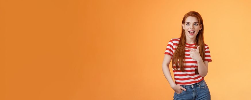 Enthusiastic amazed sassy modern redhead female attend impressive bad awesome design, look amused, observe cool place, pointing left excited, open mouth fascinated intrigued, stand orange background.