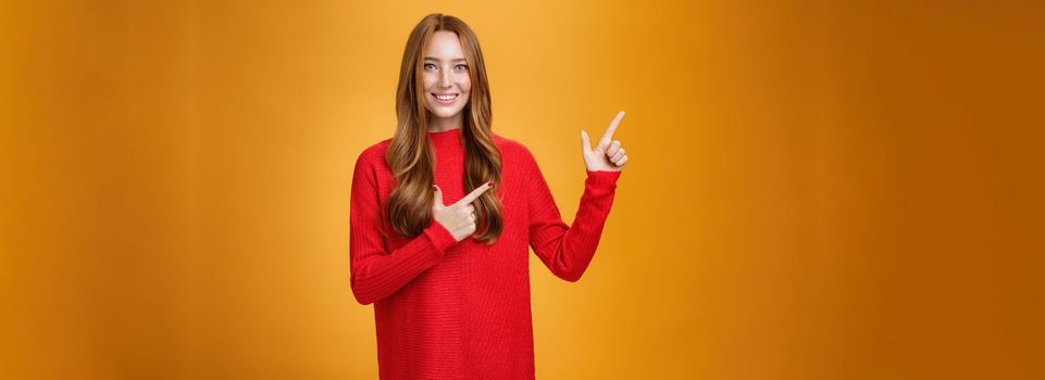 Assertive and helpful charming young female assitant in red knitted dress with ginger hair smiling friendly and pleasant as pointing at upper left corner, showing interesting thing over orange wall.