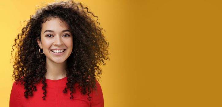 Close-up shot of pretty caucasian girl with curly hair in red dress and earrings smiling joyfully with pleased hopeful expression gazing at camera carefree, posing over yellow background.