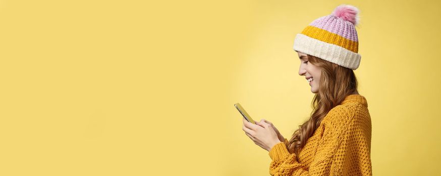 Profile shot charming outgoing relaxed young modern millennial girl texting smiling broadly holding smartphone looking display amused editing pic post online, blogger messaging followers.