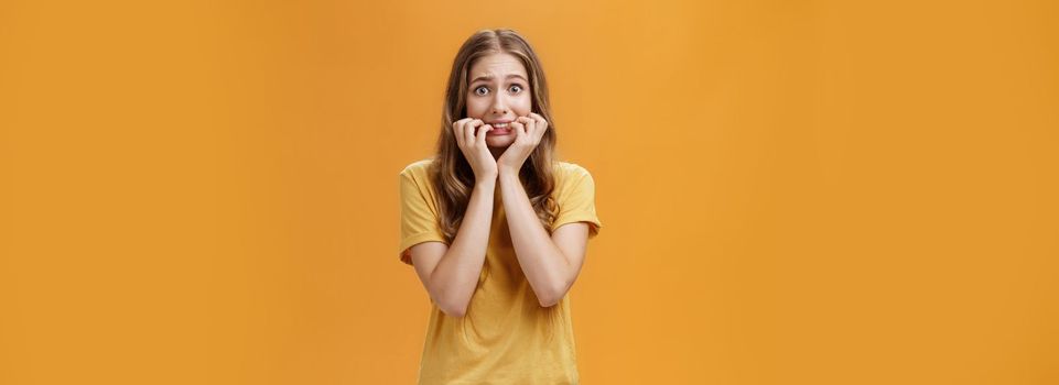 Lifestyle. Portrait of paniching nervous silly insecure young woman feeling scared trembling from fear biting fingernails keeping fingers in mouth staring at camera frightened over orange background.