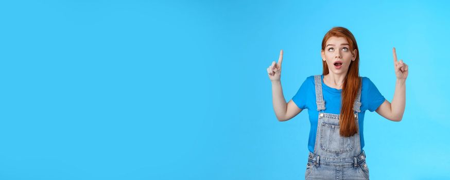 Studio shot amazed curious redhead woman drop jaw astonished, look pointing ambushed top copy space, check out up advertisement thrilled, stand surprised blue background open mouth.