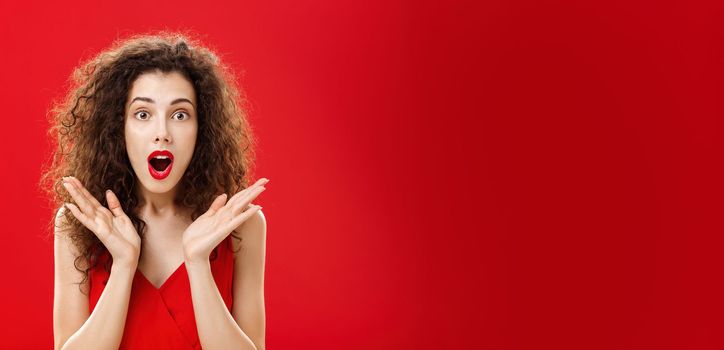 Waist-up shot of amused and astonished good-looking young elegant female. with curly hairstyle in red lipstick raising palms in surprise opening mouth and looking at camera intrigued and shocked.