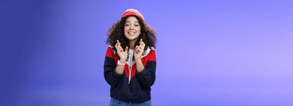 Optimistic and hopeful girl having faith in desire come true, standing with crossed fingers for good luck having wish anticipating miracle, smiling broadly at camera as wearing warm stylish red beanie.