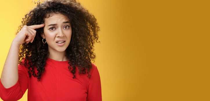 Are you crazy. Portrait of frustrated and confused woman with curly hair holding index finger near temple and raising eyebrows shocked being annoyed with stupid actions of weirdo over yellow wall.