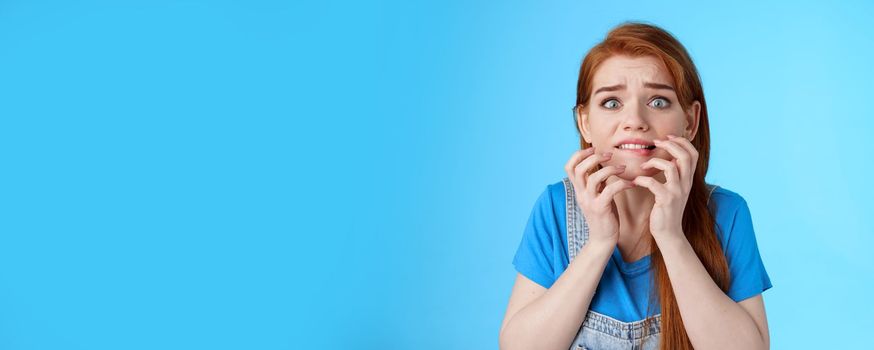Close-up intense panicking redhead woman watching scary movie feel pressured, biting lip worried, hold hands near mouth self-soothing trying calm down, stare camera anxious, blue background.