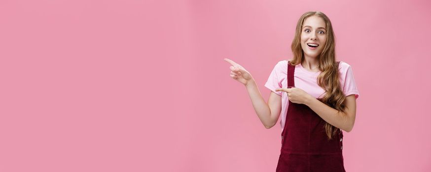 Lifestyle. Woman looking with impressed and amazed look showing cool think on left side of copy space smiling fascinated looking with admiration at camera standing delighted and surprised over pink background.