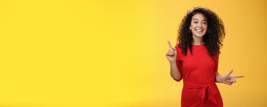 Charismatic cute and happy young curly-haired european female in red dress dancing and pointing up and right, showing sideways as presenting choices picking, making decision and smiling at camera.