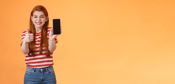 Happy satisfied redhead attractive girl cheering show thumb-up, recommend smartphone app, show mobile screen, introduce device, approve good dress show online shop cellphone, orange background.