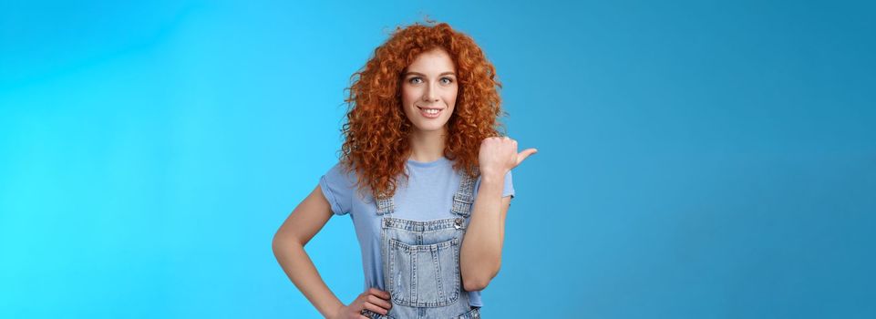 Lifestyle. Cheerful curious lively redhead good-looking curly-haired girl wearing summer overalls t-shirt pointing left thumb look camera intrigued excited explore new stores friends walking shopping mall.