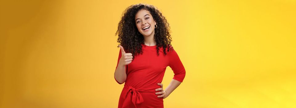 Portrait of happy optimistic young curly-haired woman in red dress laughing joyfully, showing thumbs up in approval and like gesture, delighted with awesome idea, accepting plan over yellow wall.