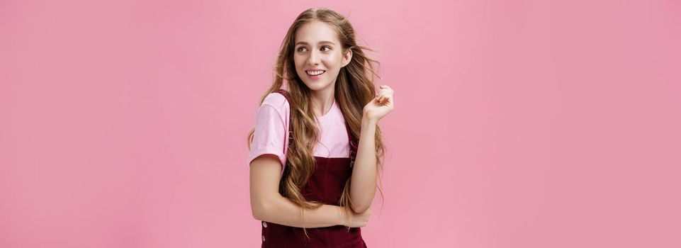 Girl looking like start from glamour magazines posing over pink background feminine and sensual looking right with flirty smile playing with hair while strands flicking in air. Fashion, style and beauty concept